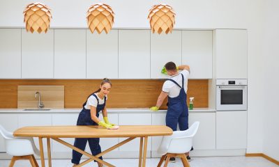 Two young janitors in blue uniform cleaning kitchen Caucasian cleaners in blue uniform wiping dust off kitchen table and cupboard. White european interior of kitchen. Cleaning service concept